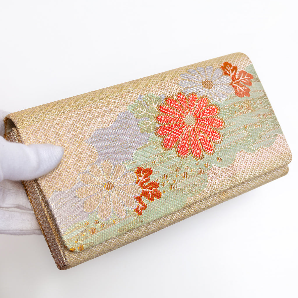 Gold Japanese silk foldover clutch with pale mint green accent and chrysanthemum motif, held in a white-gloved hand at a three-quarter angle