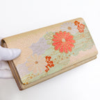 Gold Japanese silk foldover clutch with pale mint green accent and chrysanthemum motif, held in a white-gloved hand at a three-quarter angle