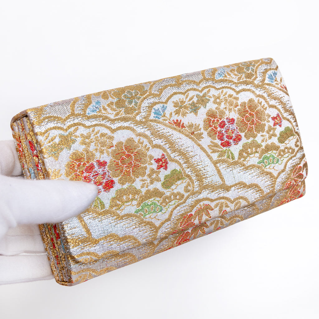 Silver and gold Japanese silk envelope clutch with intricate floral design, held in a white-gloved hand at a three-quarter angle