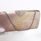 A rectangular evening clutch bag with a metallic pink and gold peacock phoenix pattern, featuring a silver rhinestone-embellished clasp and a silver chain strap, held by a gloved hand against a white background.