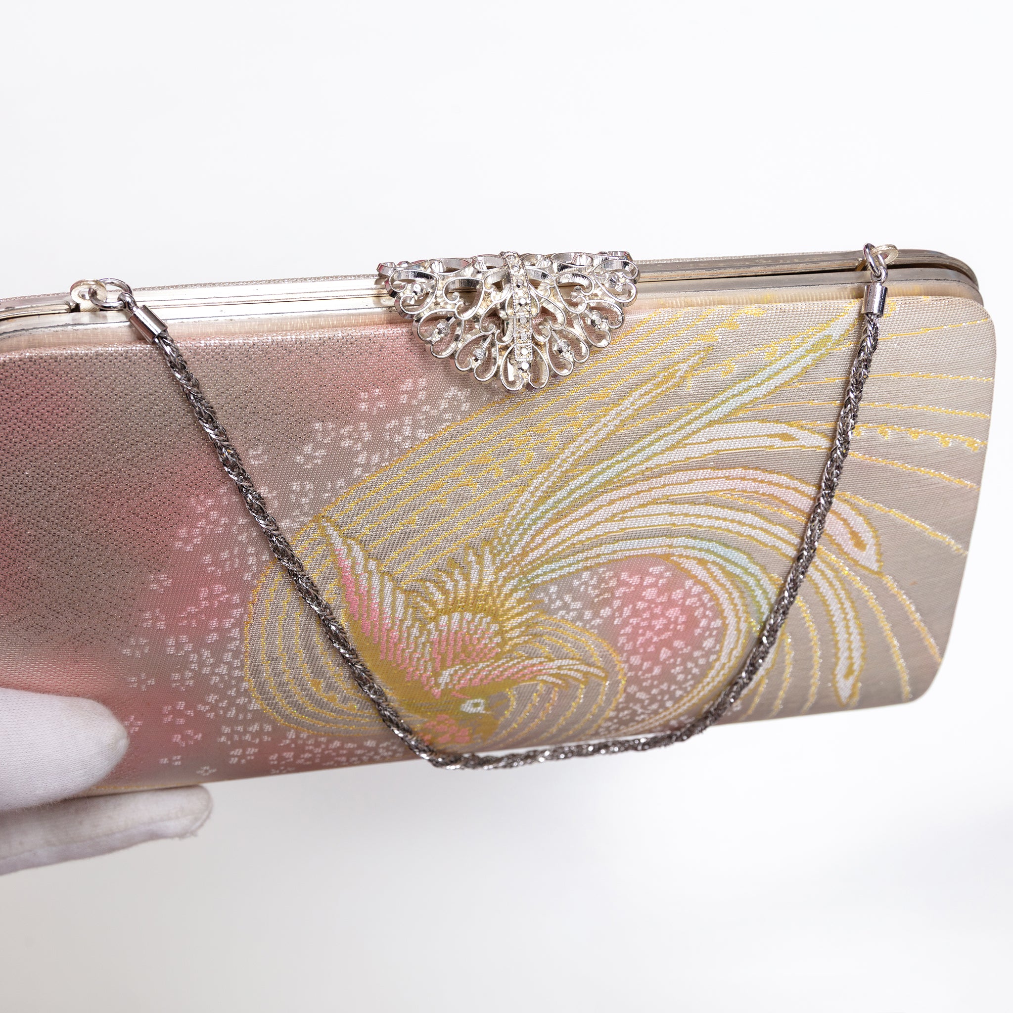 A rectangular evening clutch bag with a metallic pink and gold peacock phoenix pattern, featuring a silver rhinestone-embellished clasp and a silver chain strap, held by a gloved hand against a white background.