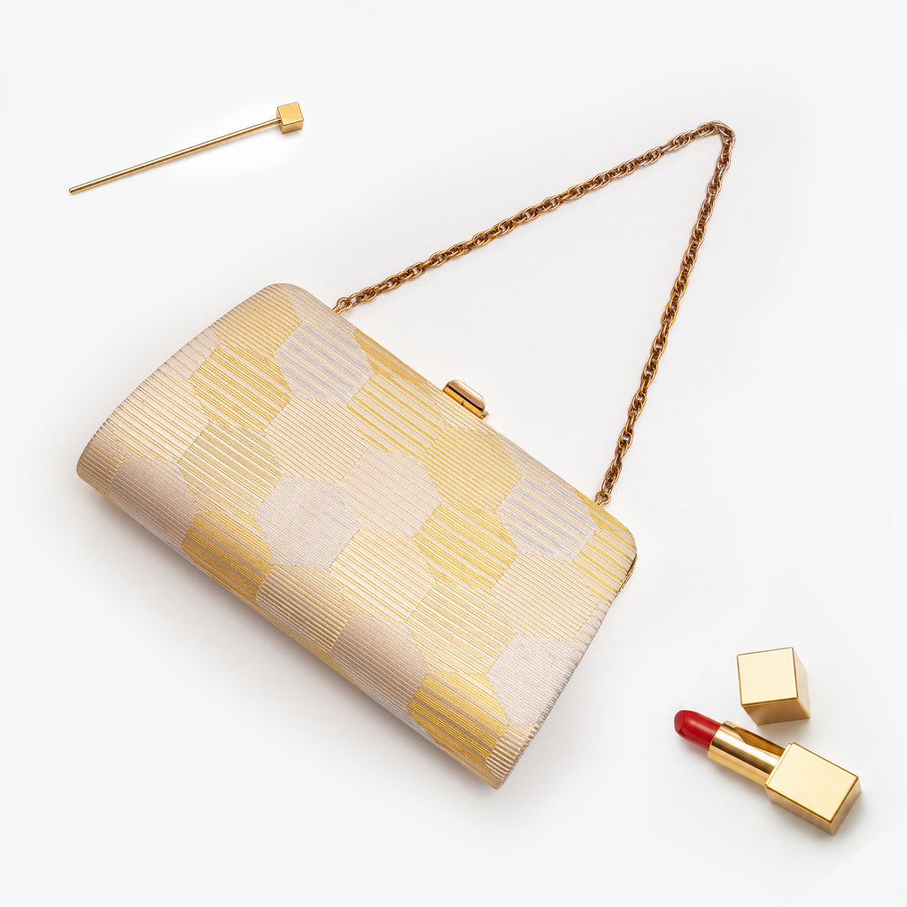 Gold and silver metallic Japanese kimono silk clutch bag with geometric fortune hexagon pattern, a gold chain strap, and a gold clasp, styled on a white background with a gold pin, lipstick, and lipstick case.