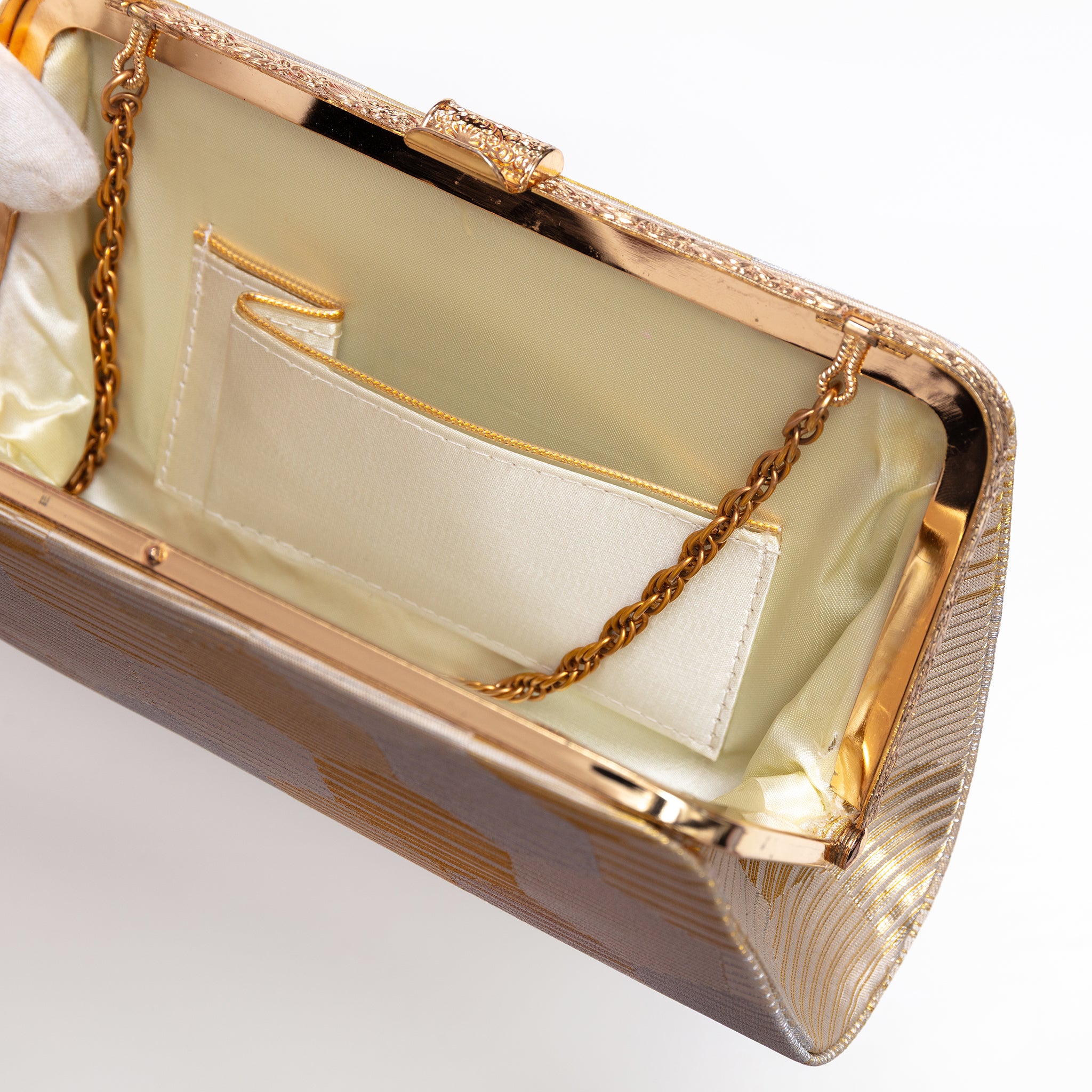 Close-up of the interior of a metallic gold evening clutch bag with a gold chain strap and a cream satin lining, shown on a white background.