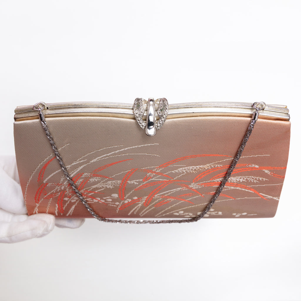 A vintage-style evening clutch bag with a metallic gold and rose gold satin body, decorated with orange and silver floral patterns, featuring a silver-tone metal clasp and a silver chain strap. A white-gloved hand is visible on the left holding the bag.