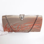 A vintage-style evening clutch bag with a metallic gold and rose gold satin body, decorated with orange and silver floral patterns, featuring a silver-tone metal clasp and a silver chain strap. A white-gloved hand is visible on the left holding the bag.