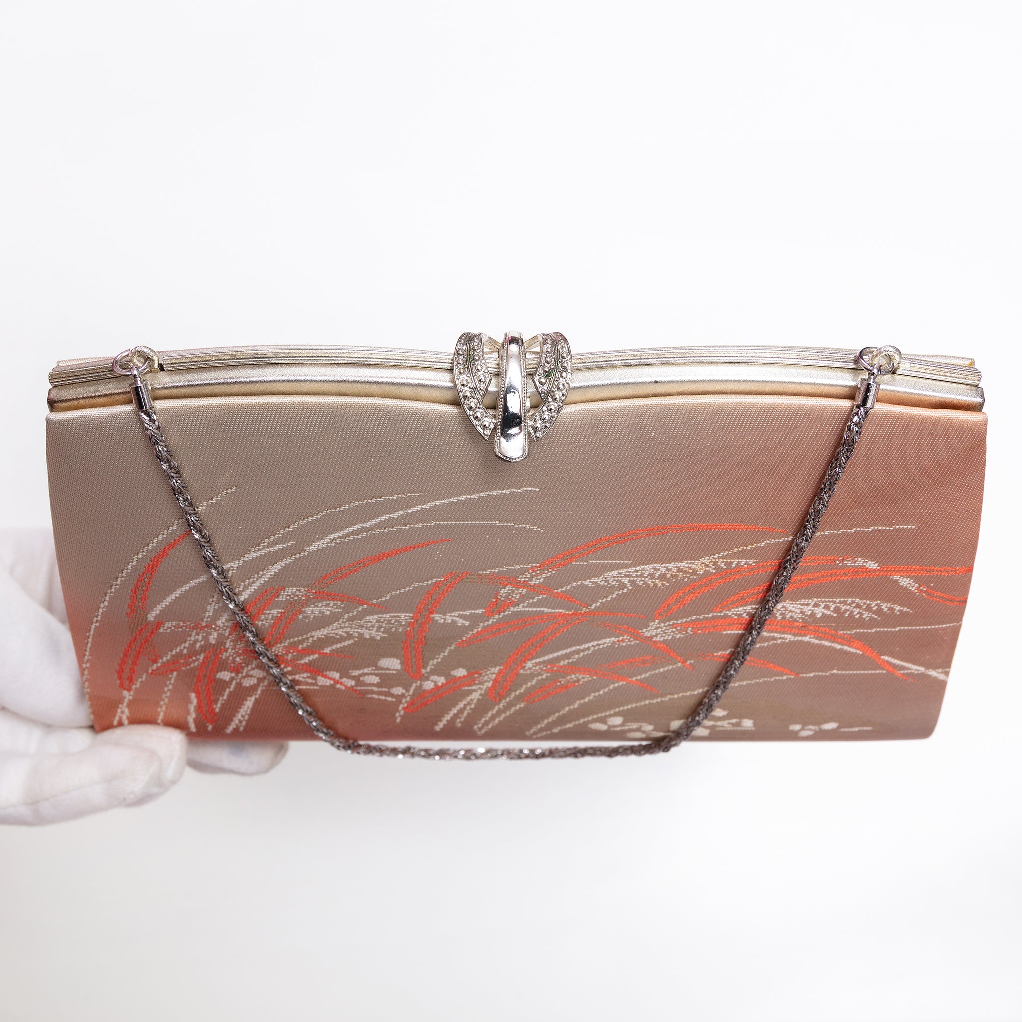 A vintage-style evening clutch bag with a metallic gold and rose gold satin body, decorated with orange and silver floral patterns, featuring a silver-tone metal clasp and a silver chain strap. A white-gloved hand is visible on the left holding the bag.