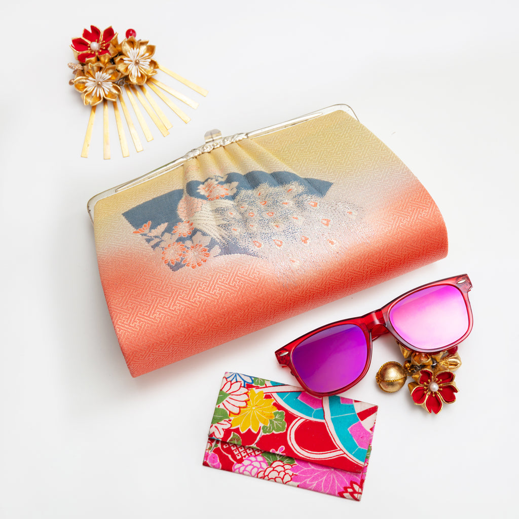 Peach and yellow vintage Japanese silk clutch bag with peacock and fan design on a white surface surrounded by accessories and sunglasses.
