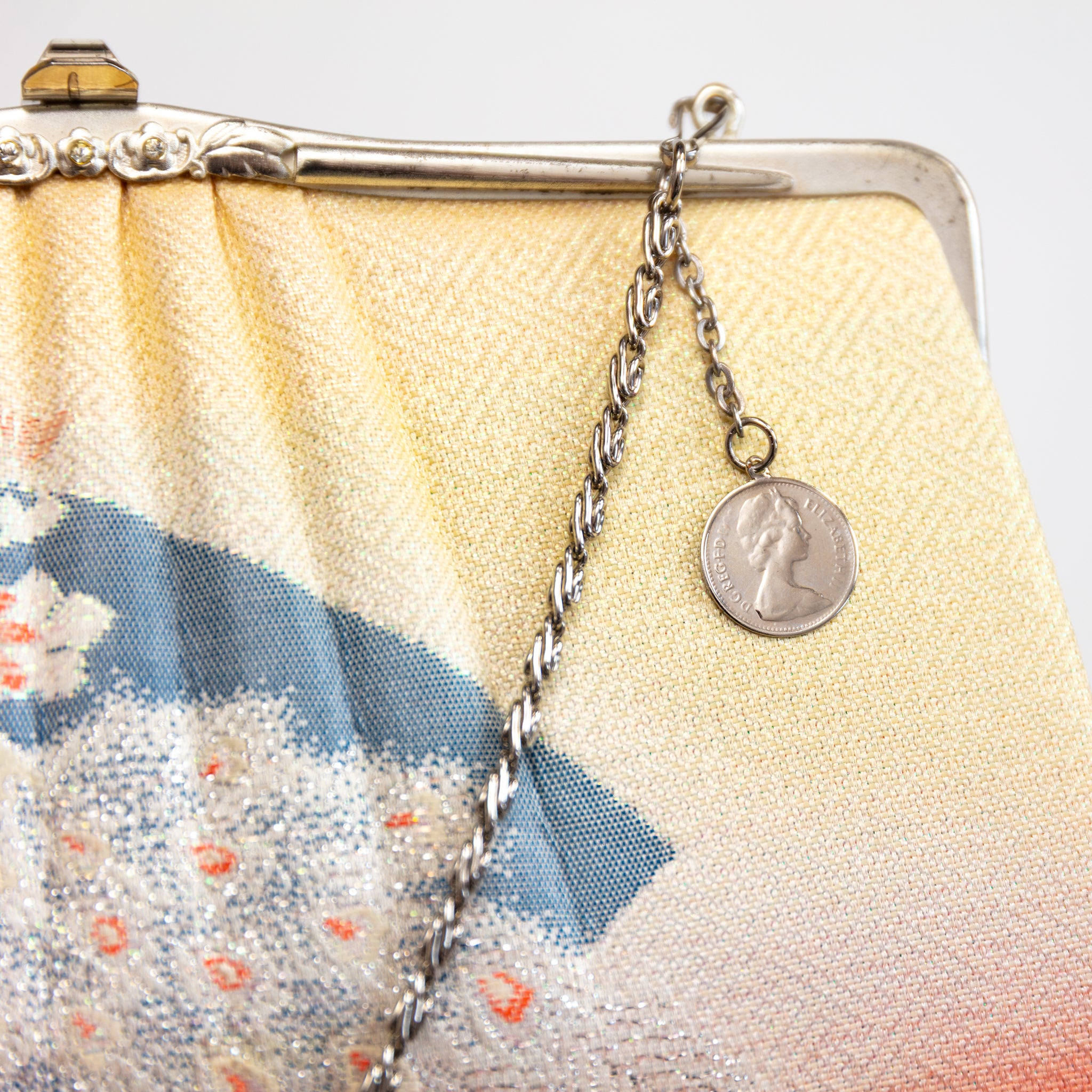 Close up detail of a vintage Japanese silk clutch bag corner showing the silver bag frame, floral metal clasp, silver fancy chain strap and English coin charm draped over the yellow silk front of the bag.