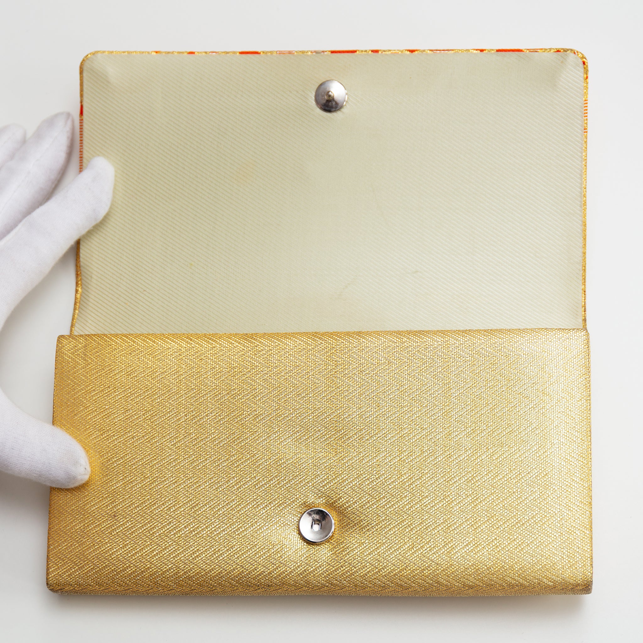 Gold clutch with textured surface and silver buttons held by a white-gloved hand on a light background
