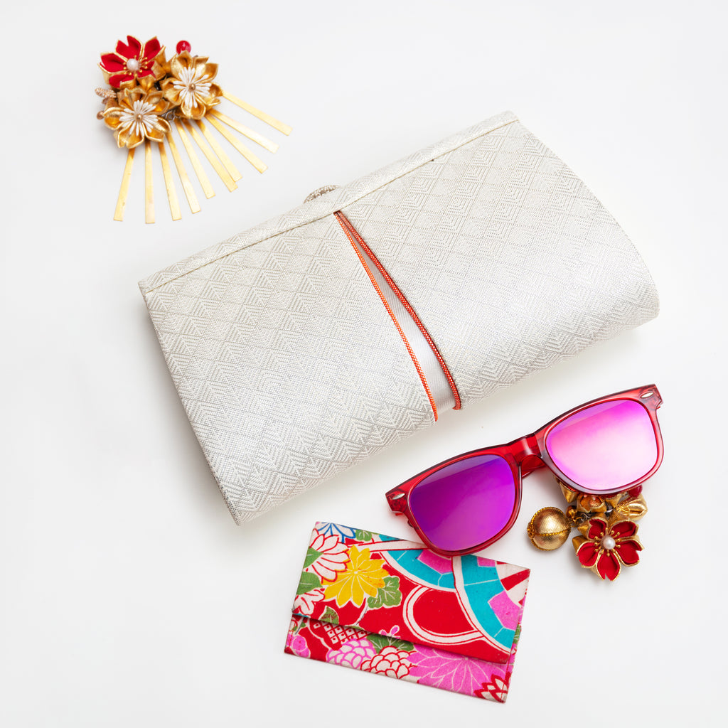 Vintage Japanese silk clutch bag in white and silver with orange accent trim surrounded y accessories on white background.