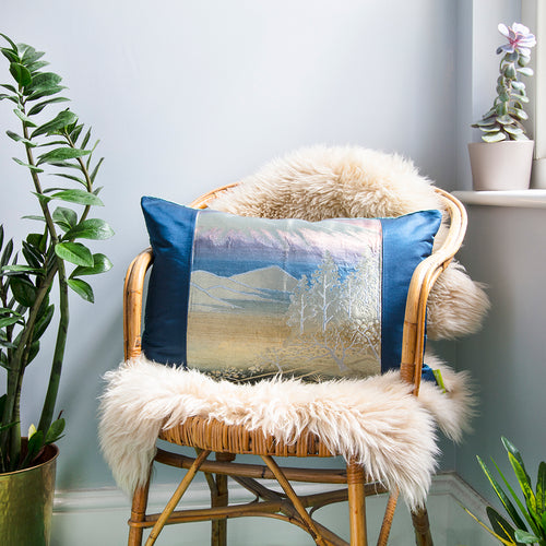 Silver and blue metallic mountain scene cushion on a wicker chair in a grey room with plants.
