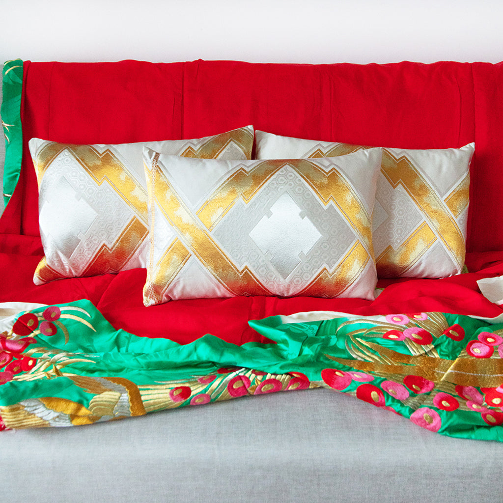 Set of three patterned obi cushions in gold, cream and silver sitting on a neutral sofa that has a red and turquoise kimono silk throw over it.