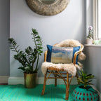 View of a blue and silver obi cushion on a chair in a room with plants, mirror and turquoise rug.