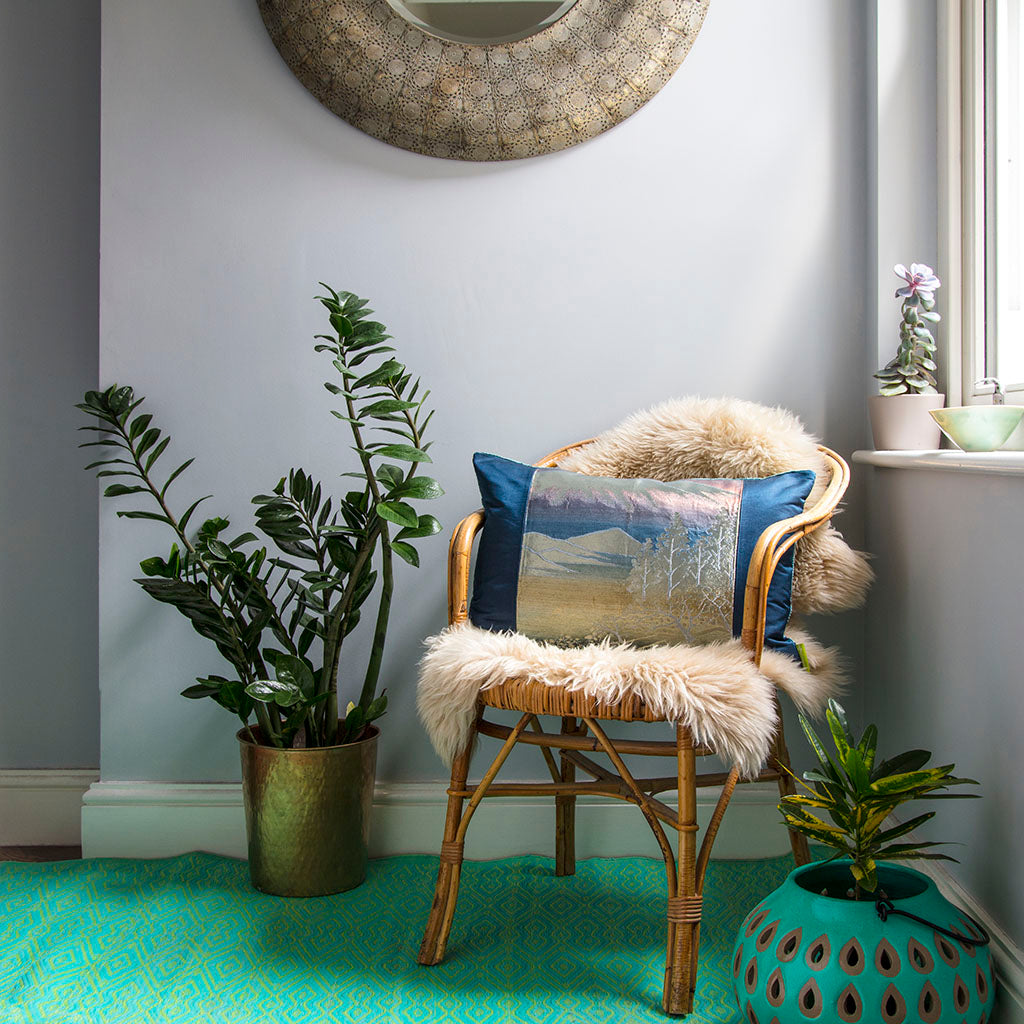 View of a blue and silver obi cushion on a chair in a room with plants, mirror and turquoise rug.