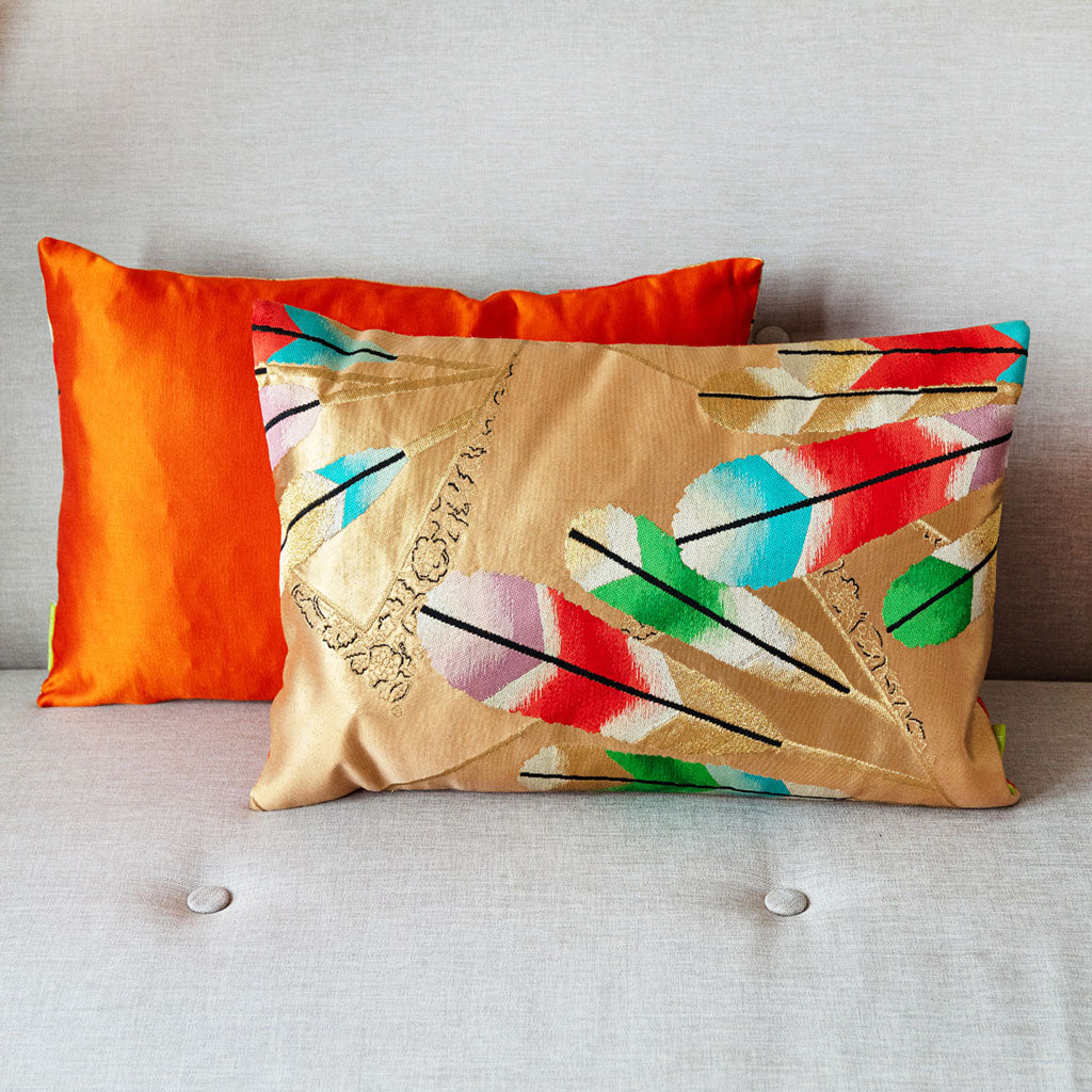 Gold obi cushion with feather motifs in front of an orange silk cushion (the colour of the reverse of the cushion) on a neutral sofa.