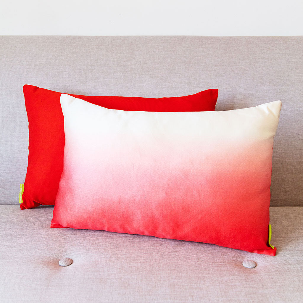 Rectangular pink to white ombre cushion on a neutral sofa with a red cushion behind it showing the colour of the reverse.