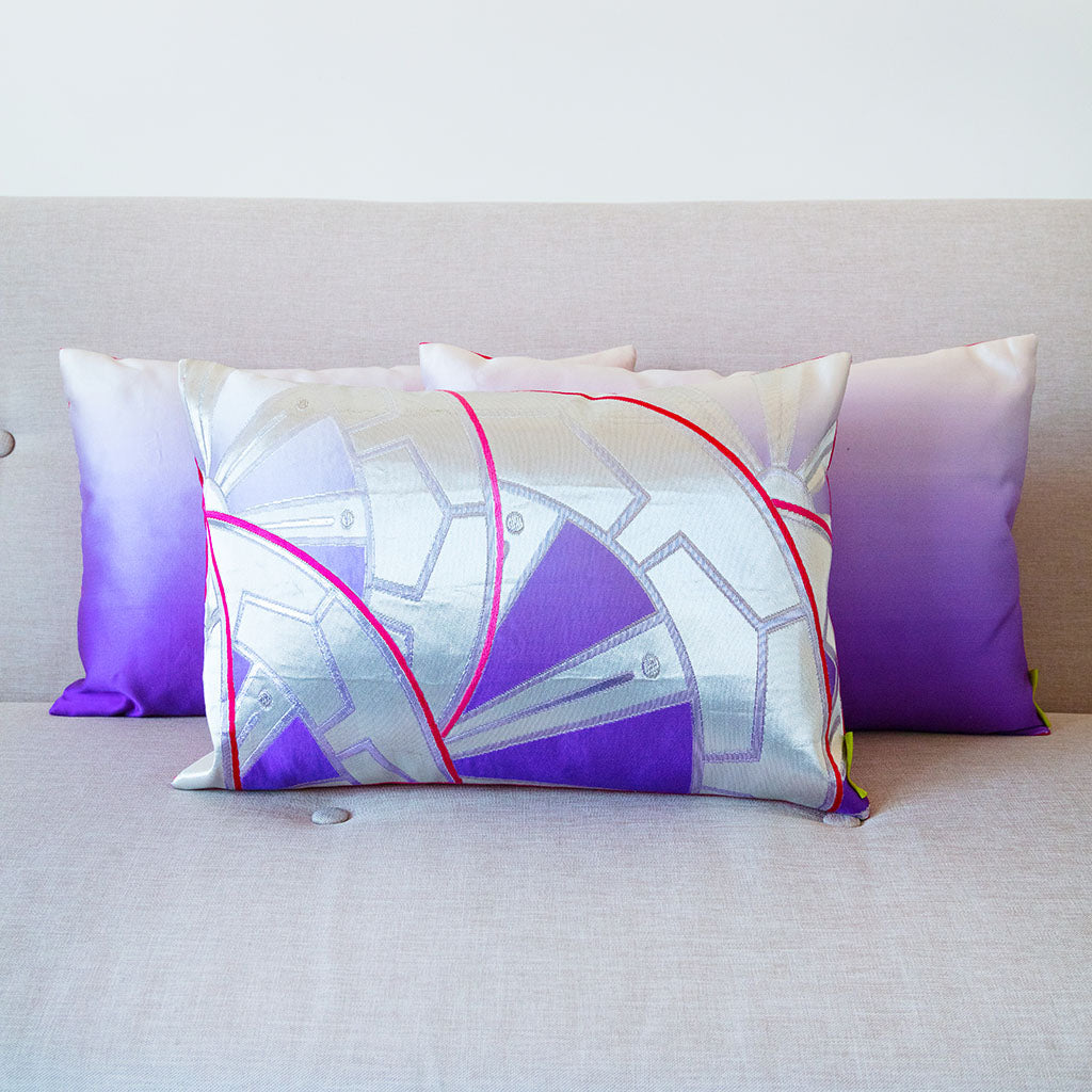 Set of three purple to white ombre rectangular cushions on a neutral sofa. Patterned cushion at the front with silver and pink accents, and 2 plain ombre cushions behind.