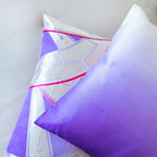 View of two cushion edges, the top cushion is ombre purple to white silk and the cushion below has genji wheel pattern in silver with pink accents.